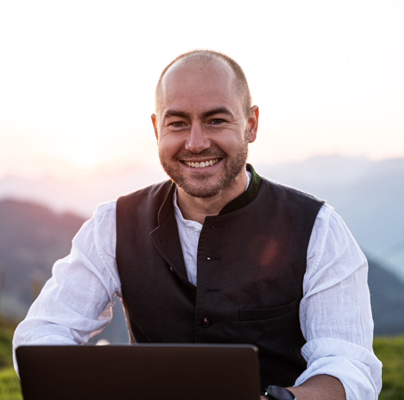 Portraitaufnahme von Benedikt Holzmann, breit grinsend an einem Tisch in den Bergen sitzend, bei angenehmer Sonneneinstrahlung